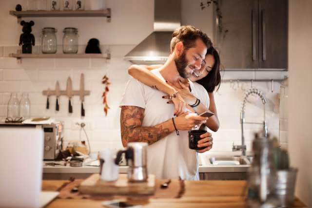 Couple embracing in a modern kitchen, smiling and looking at a phone.