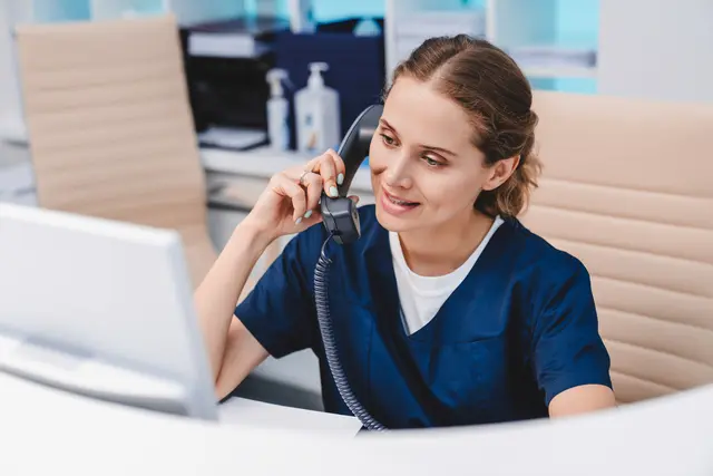 Nurse on phone at computer, wearing blue scrubs, smiling.
