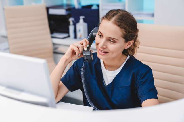 Nurse on phone at computer, wearing blue scrubs, smiling.