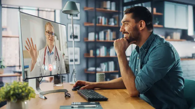 Man in a video call with a doctor on a computer, smiling in a modern home office.