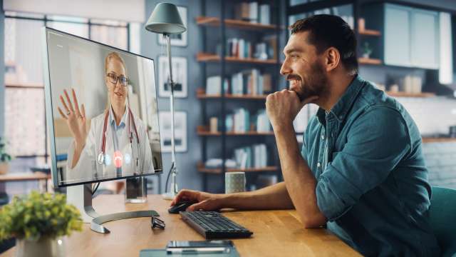 Man in a video call with a doctor on a computer, smiling in a modern home office.