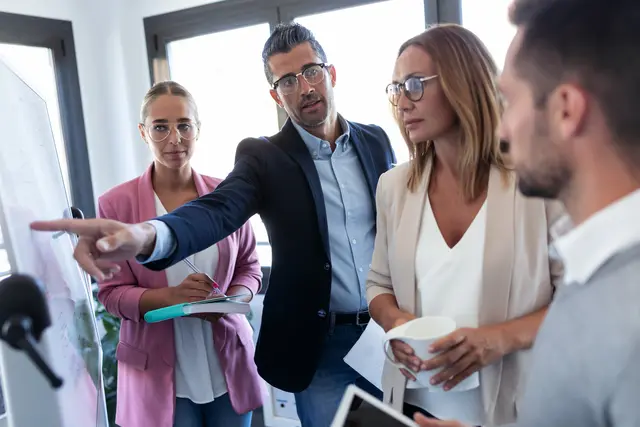 Group of colleagues in a meeting, one pointing at a whiteboard.