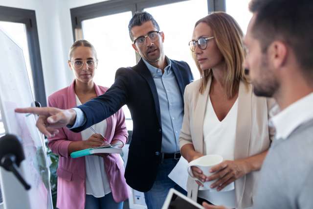 Group of colleagues in a meeting, one pointing at a whiteboard.